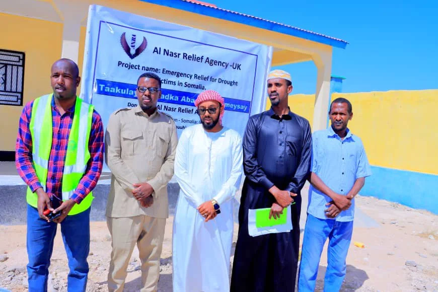 Five men standing in front of a banner at an outdoor site, with a yellow and blue building in the background. The banner indicates a relief project for drought victims in Somalia, organized by Al Nasr Relief Agency - UK.