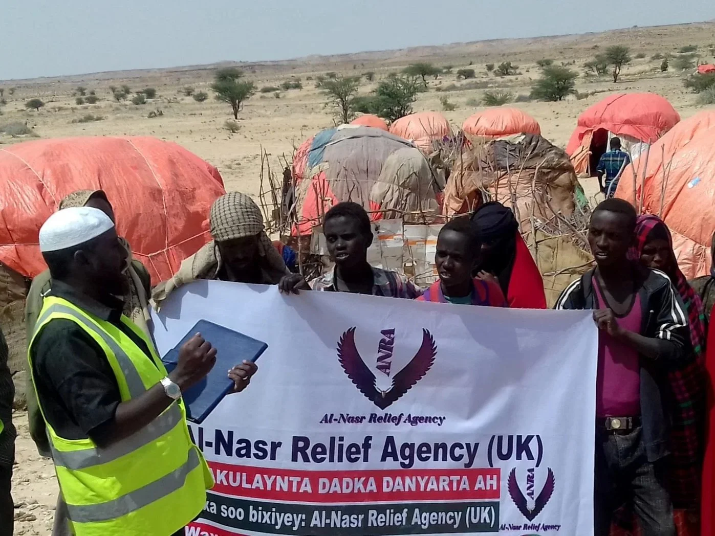 Group of people holding a banner with the text 'Al-Nasr Relief Agency (UK)' in a desert setting, with tents in the background.