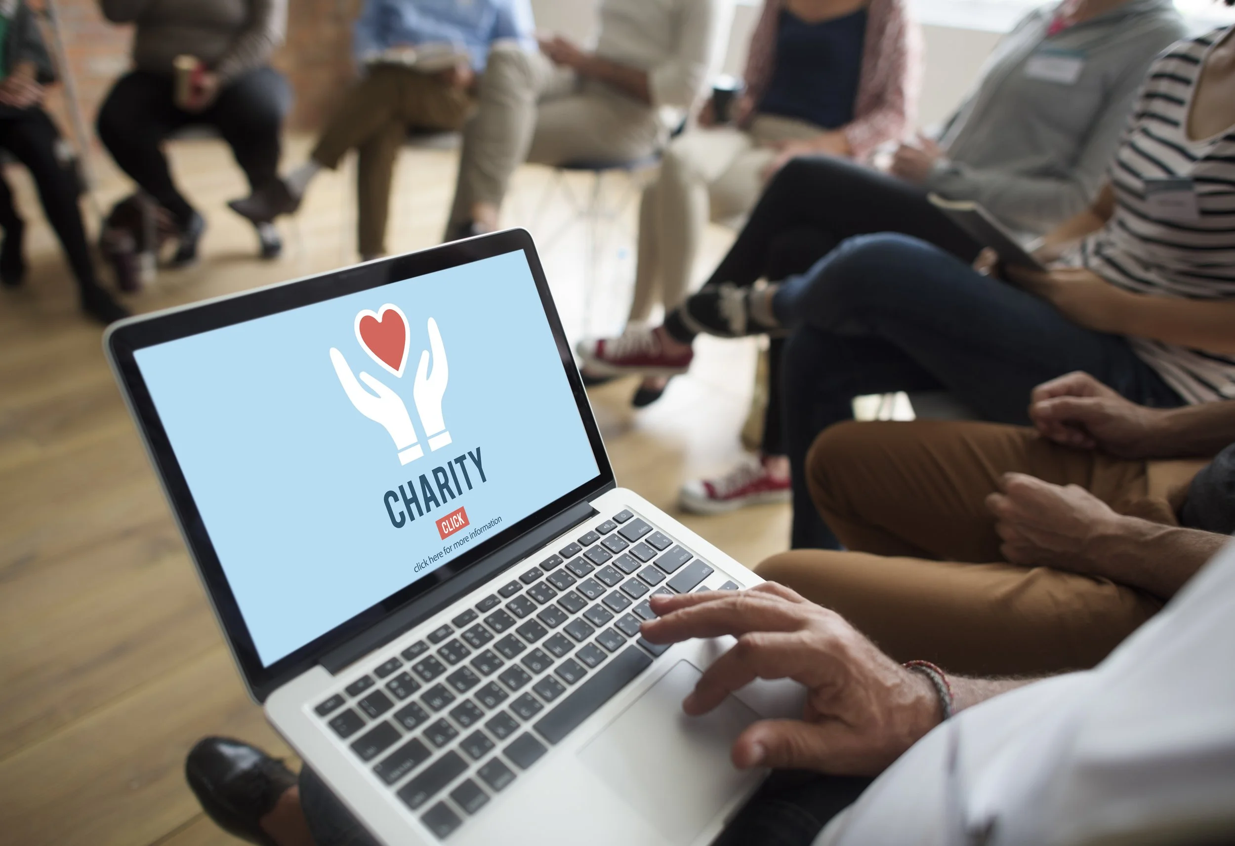 Close-up of a laptop screen displaying a charity donation webpage, with a group of people sitting in the background at a community event.