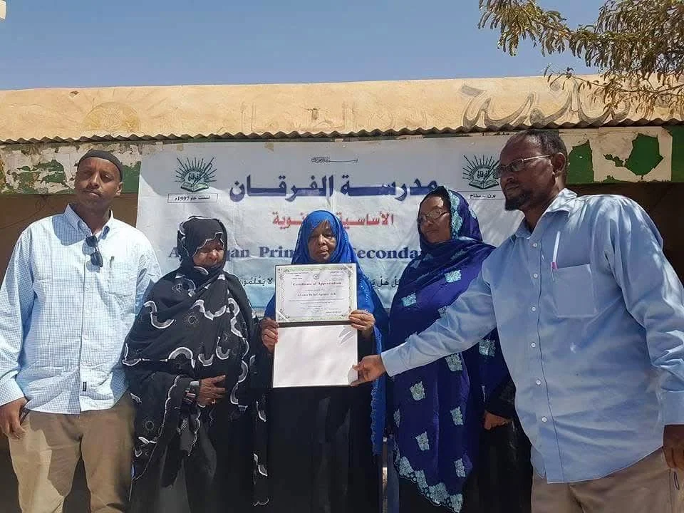 Group of five adults, including women in hijabs, standing outdoors in front of a banner with Arabic and English text. One woman is holding a certificate, and a man on the right touches it, while others look on.