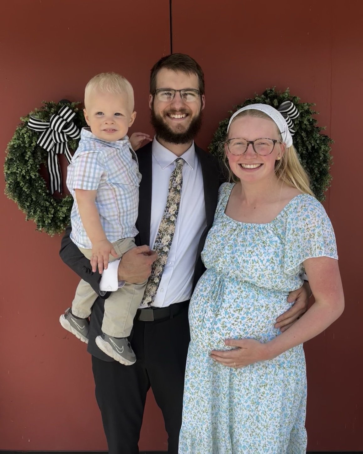 A family of three standing in front of a red wall with holiday wreaths, smiling at the camera. The man is holding a young boy, and a pregnant woman is standing on the right.
