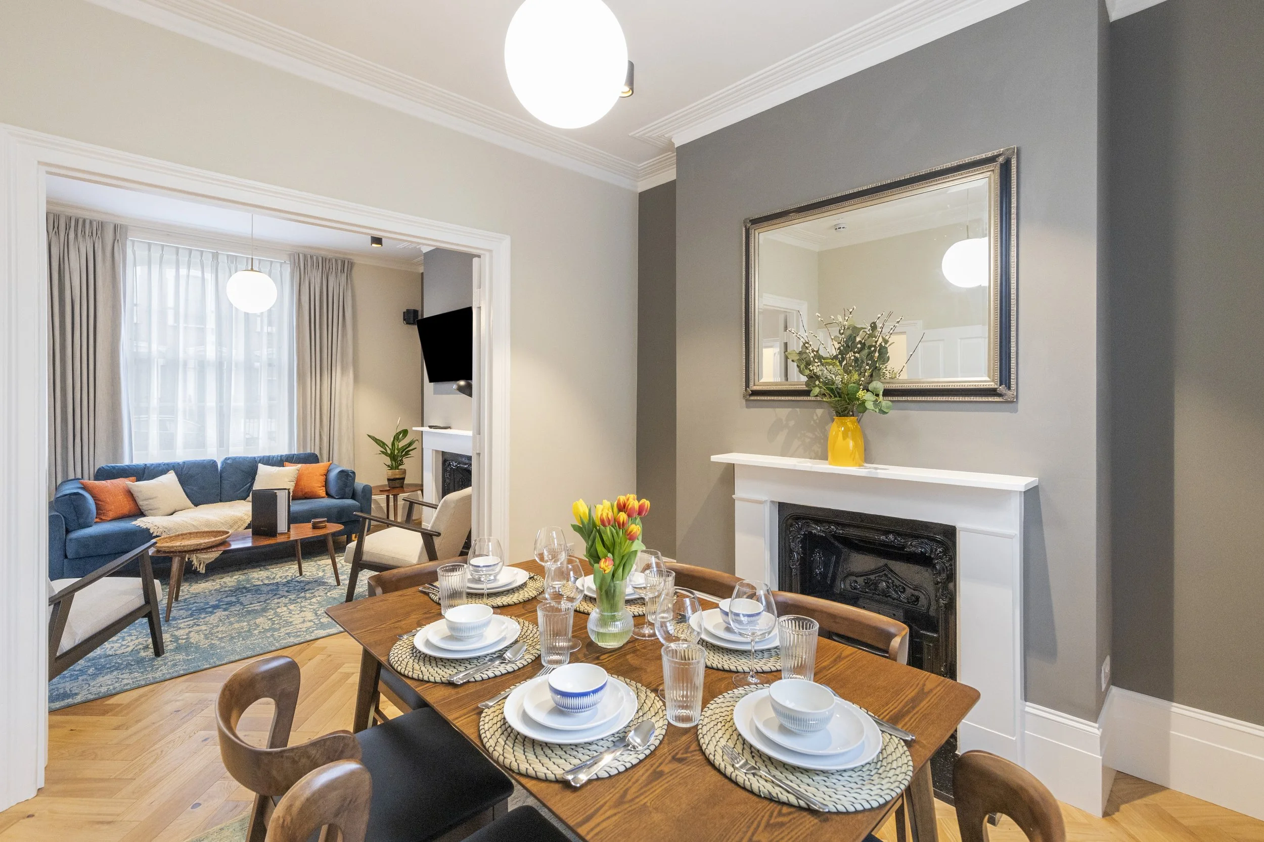 A living and dining room with a wooden dining table set with plates, glasses, and utensils, a blue sofa with orange and white cushions, a window with beige curtains, a mirror above a white mantelpiece with a yellow vase and green plants, and a flat-screen TV in the corner.