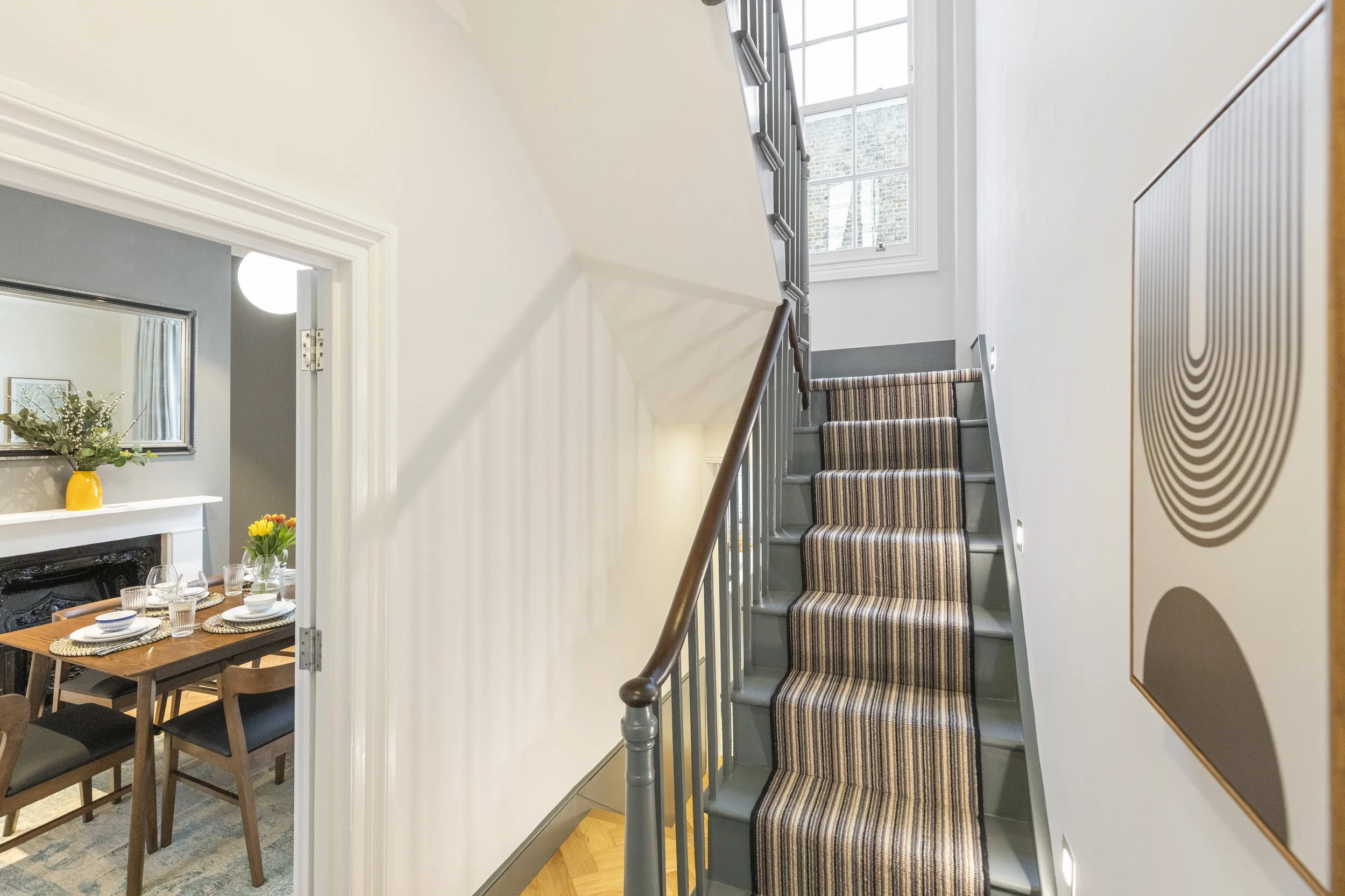 Interior view of a staircase with striped carpet runner, gray risers, and white walls, next to a window letting in natural light.