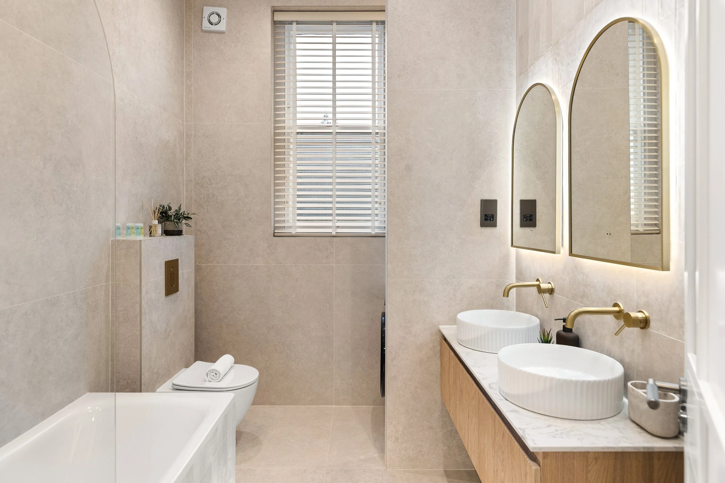 Modern bathroom with beige tiled walls and floor, a window with white blinds, a built-in bathtub, a floating vanity with dual round sinks, gold fixtures, and a mirror, along with decorative plants and toiletries.