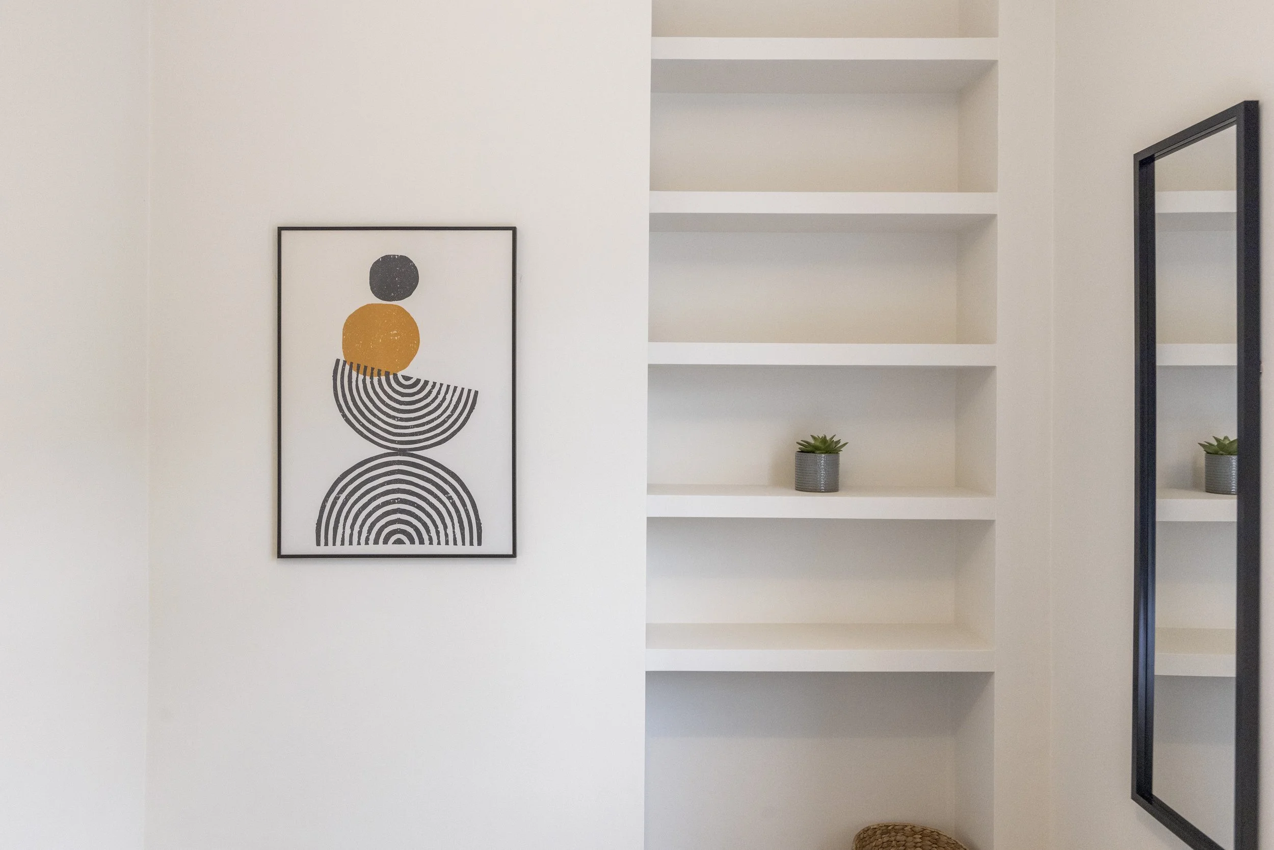 Empty white bookshelf with three shelves, two potted plants on the bottom shelf, a black framed abstract art print on the wall to the left, and a black rectangular mirror on the right.