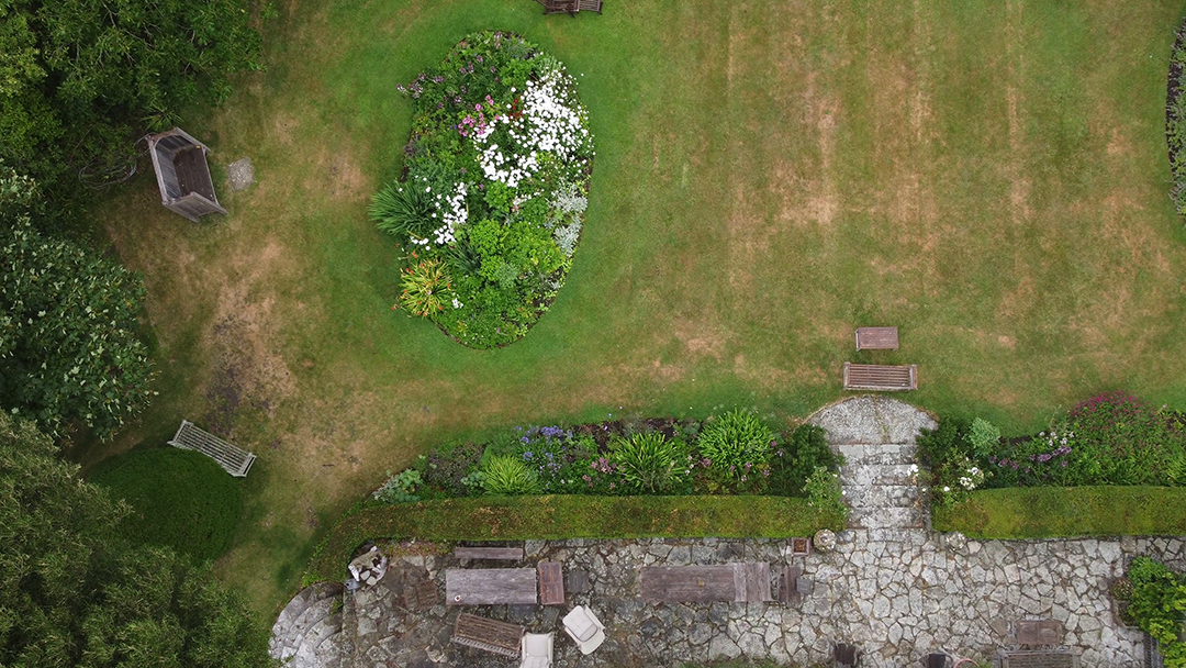 A bird's eye view of a garden with a stone pathway, flower beds, benches, a small shed, and well-maintained grass