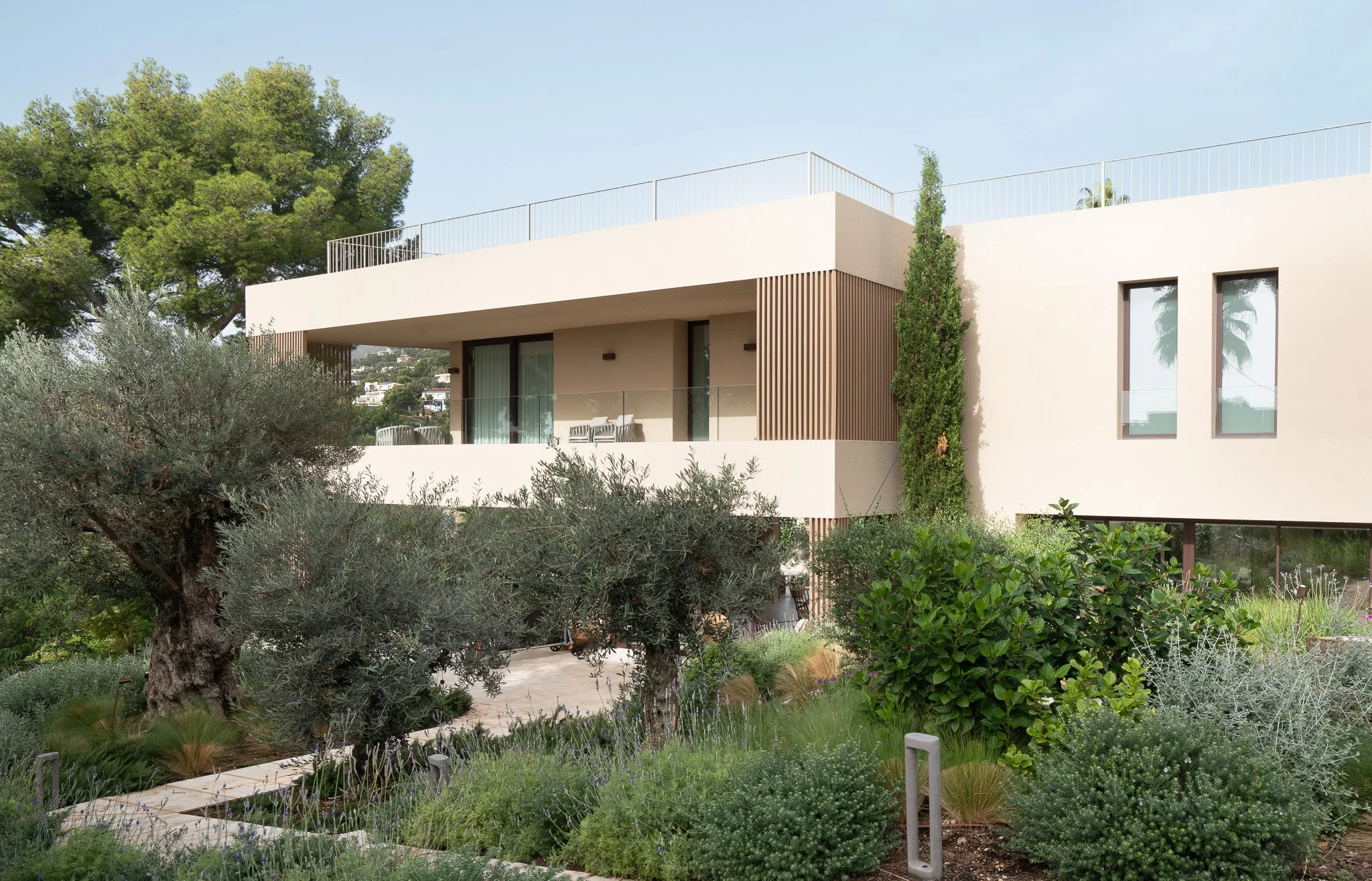 Modern house with white exterior, large glass doors, and balcony, surrounded by trees and lush garden.