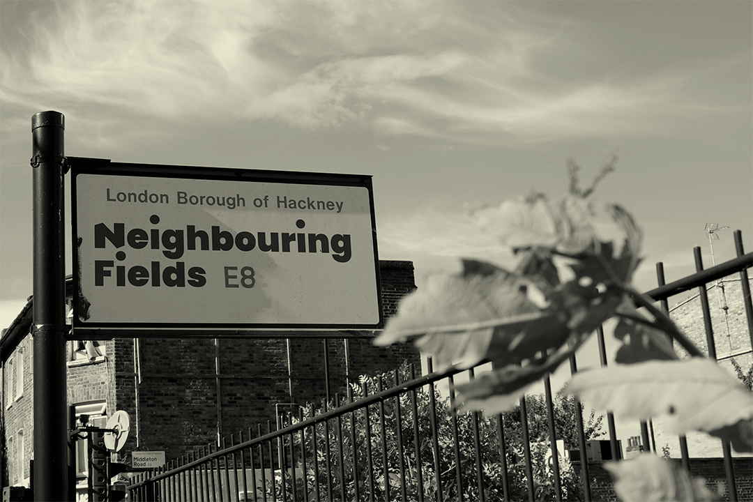 Sign for Neighbouring Fields in London Borough of Hackney, with leaves and a fence in the foreground and a building and cloudy sky in the background.