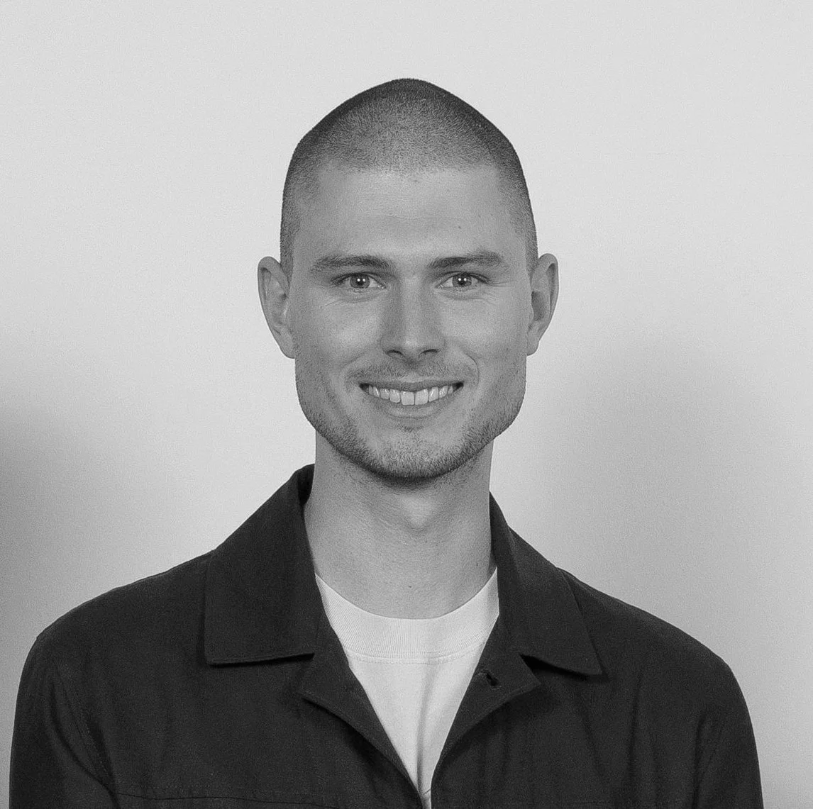 Black and white portrait of a young man with short hair, smiling, wearing a collared shirt and a t-shirt underneath, against a plain background.