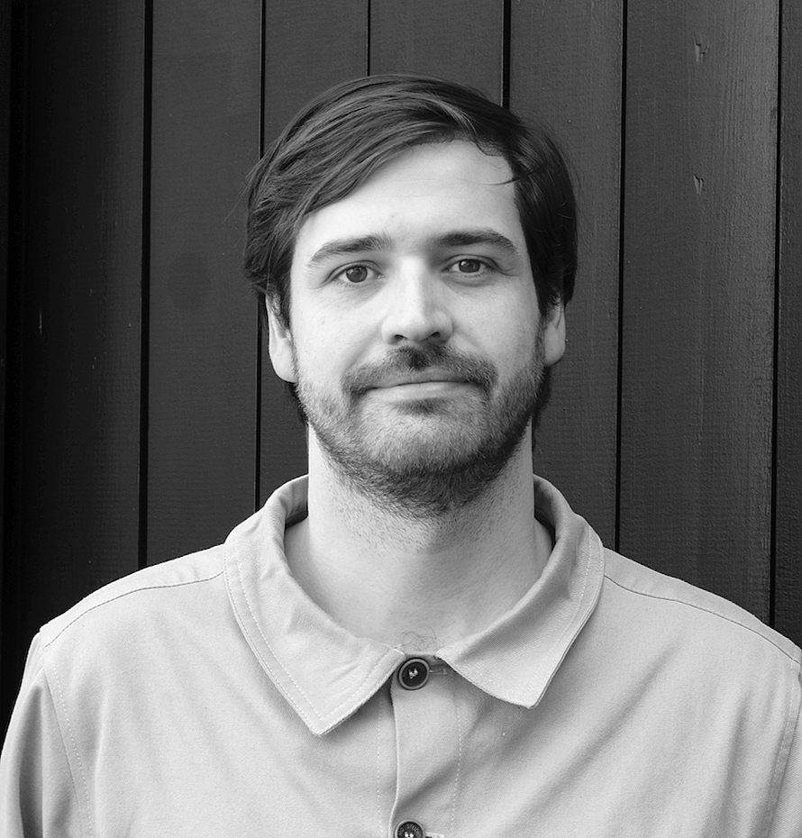 Black and white portrait of a young man with medium-length hair, beard, wearing a collared shirt, standing in front of dark wooden paneling.
