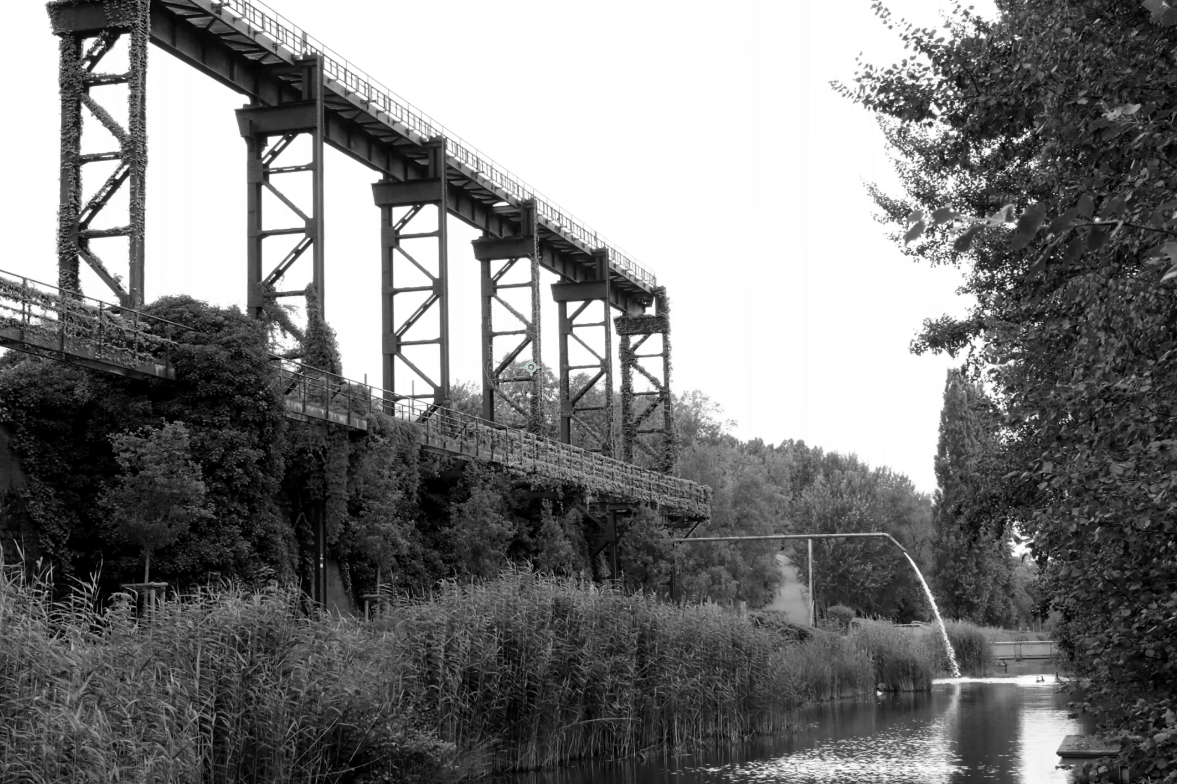 Black and white photo of a large industrial bridge over a canal with water flowing from a pipe on the right, surrounded by trees and bushes.