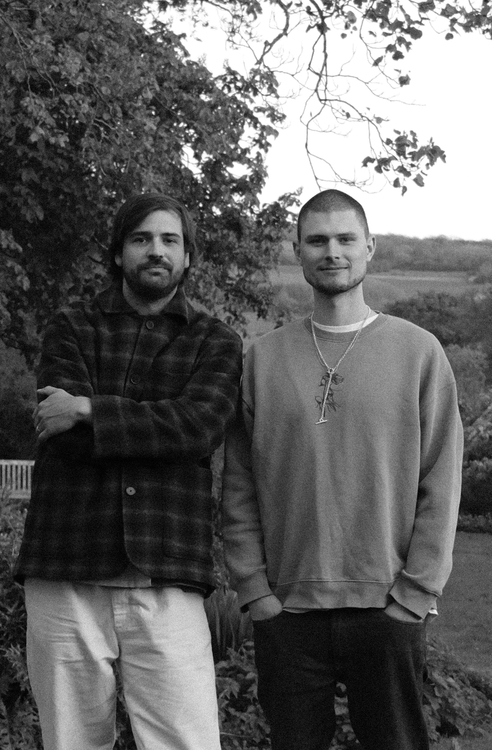Two young men standing outdoors in front of trees, smiling at the camera, in black and white.