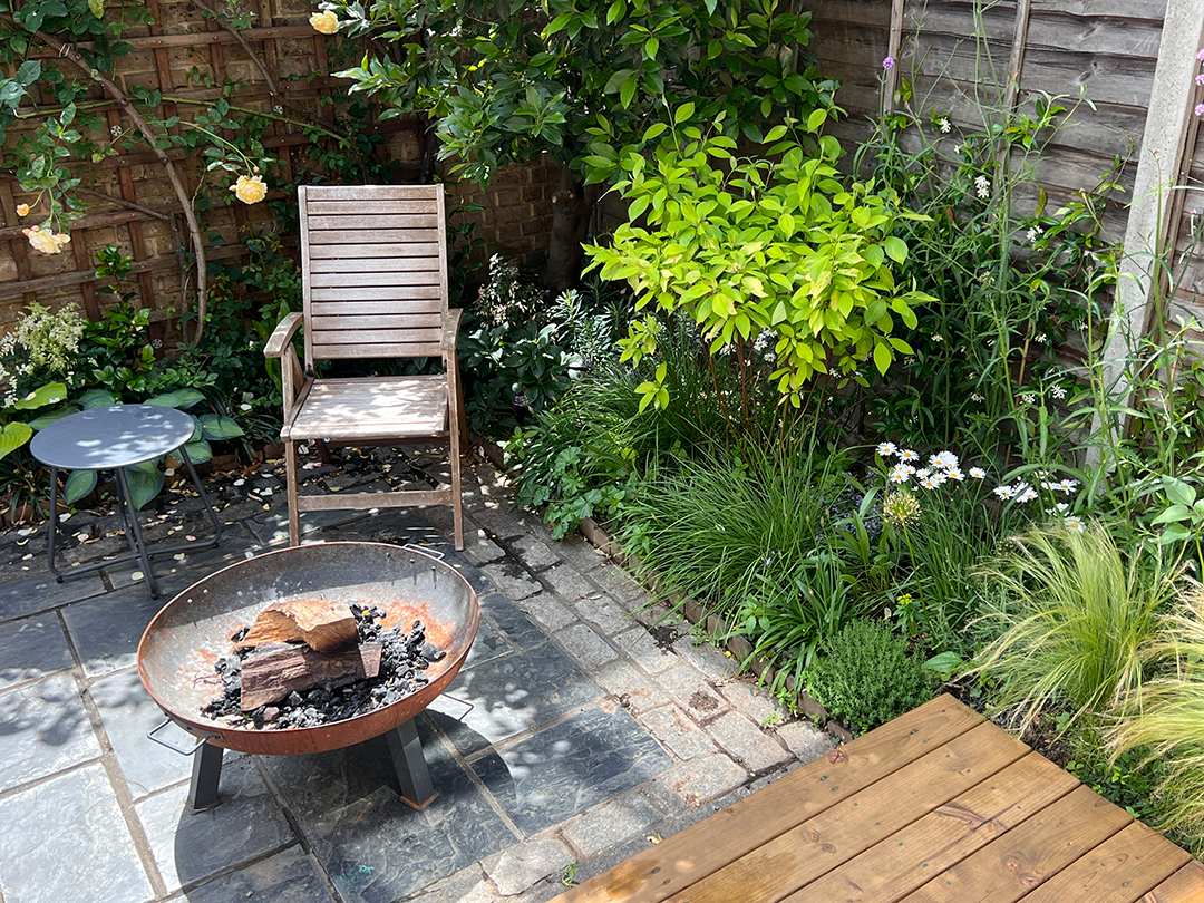A small outdoor patio area with a fire pit, wooden chair, small side table, and lush green plants and flowers along a wooden fence.