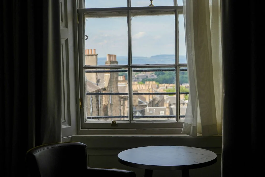 View through a window of buildings, rooftops, and distant hills on a cloudy day, with sheer curtains partially drawn.