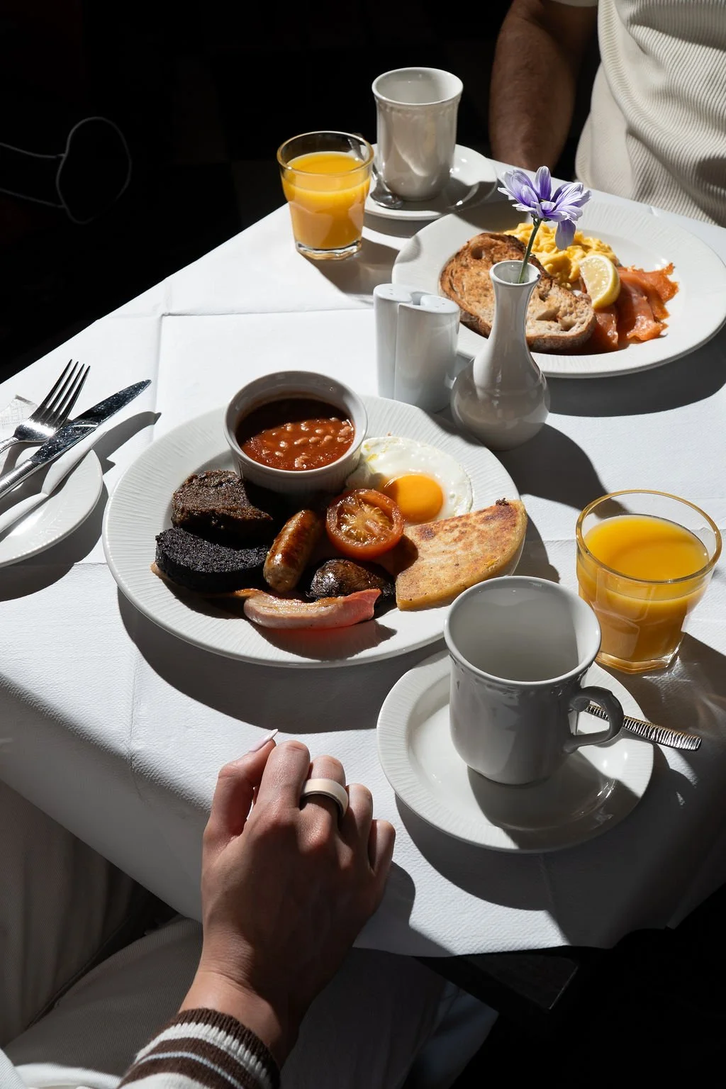A breakfast table set with plates of cooked breakfast foods, cups of coffee, glasses of orange juice, and a hand reaching for a mug.