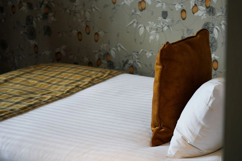 A neatly made bed with white sheets, a plaid blanket, and a large brown headboard against a patterned wallpaper with orange fruit motifs.
