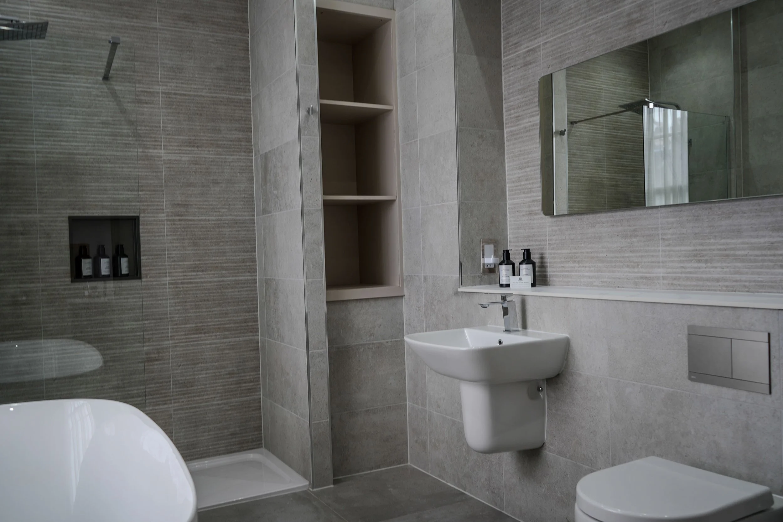 Modern bathroom with tiled walls and floor, a wall-mounted sink with soap bottles, a large mirror, an open shelving niche, and a shower area with a glass door.