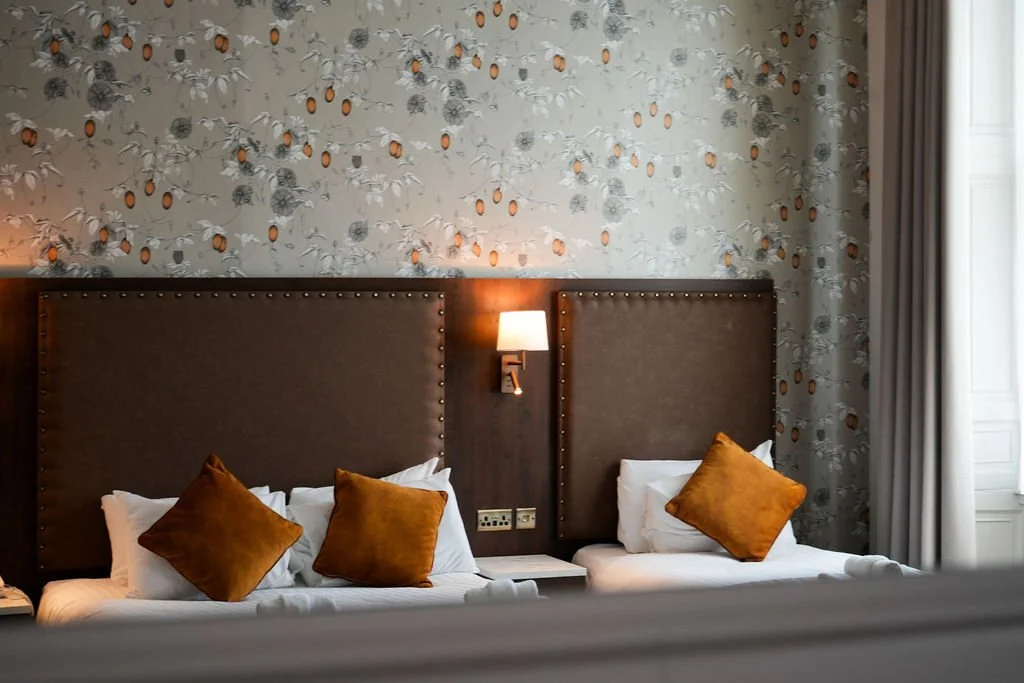 Hotel bedroom with two beds, each with white linens and rust-colored pillows, large padded headboards, floral wallpaper featuring fruits and leaves, a wall-mounted lamp, electrical outlets, and a window with curtains.
