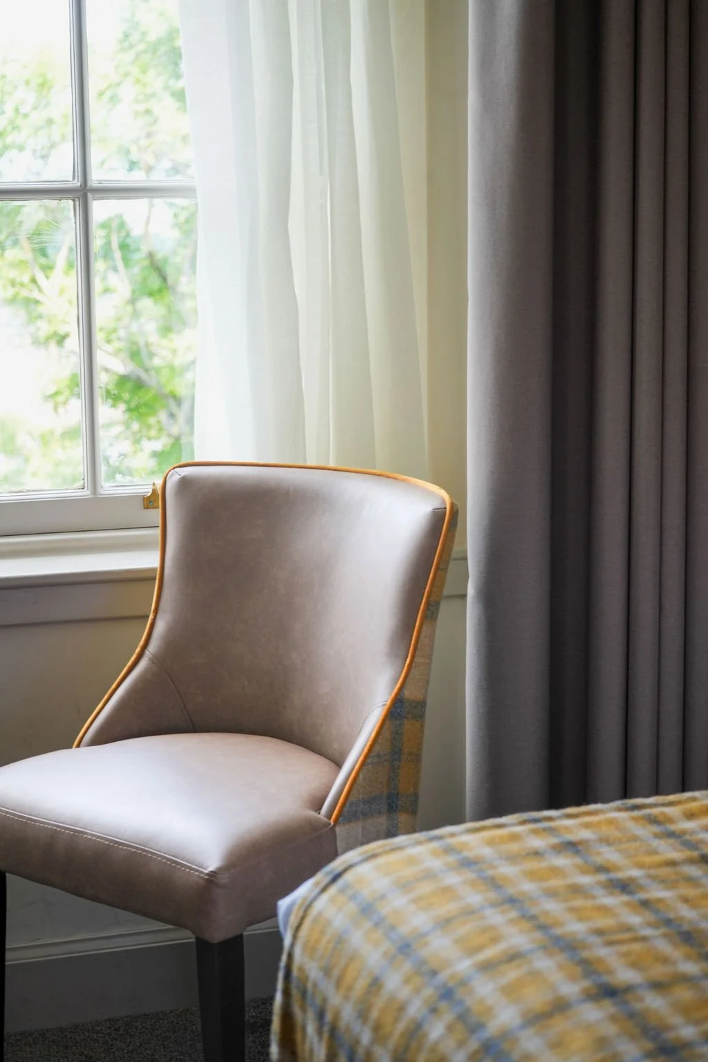 A beige upholstered armchair with a wooden frame next to a window with white and grey curtains, and a bed with yellow checkered bedding in the foreground.