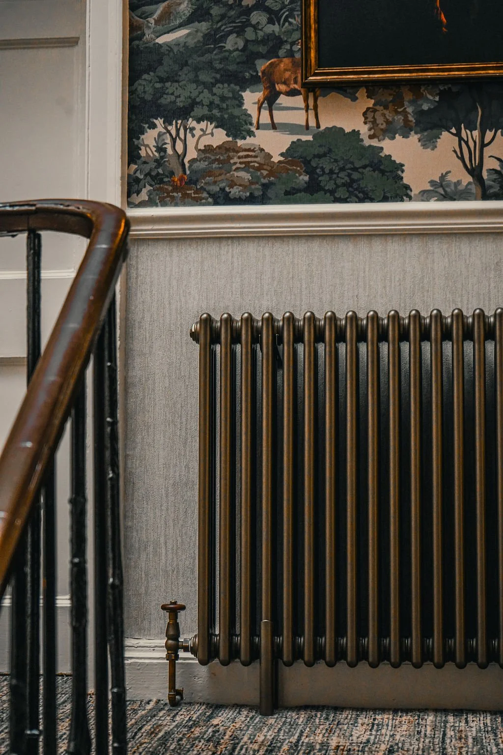 A bronze radiator against a textured wall in a room with a patterned carpet and a framed landscape painting of trees and a dog.