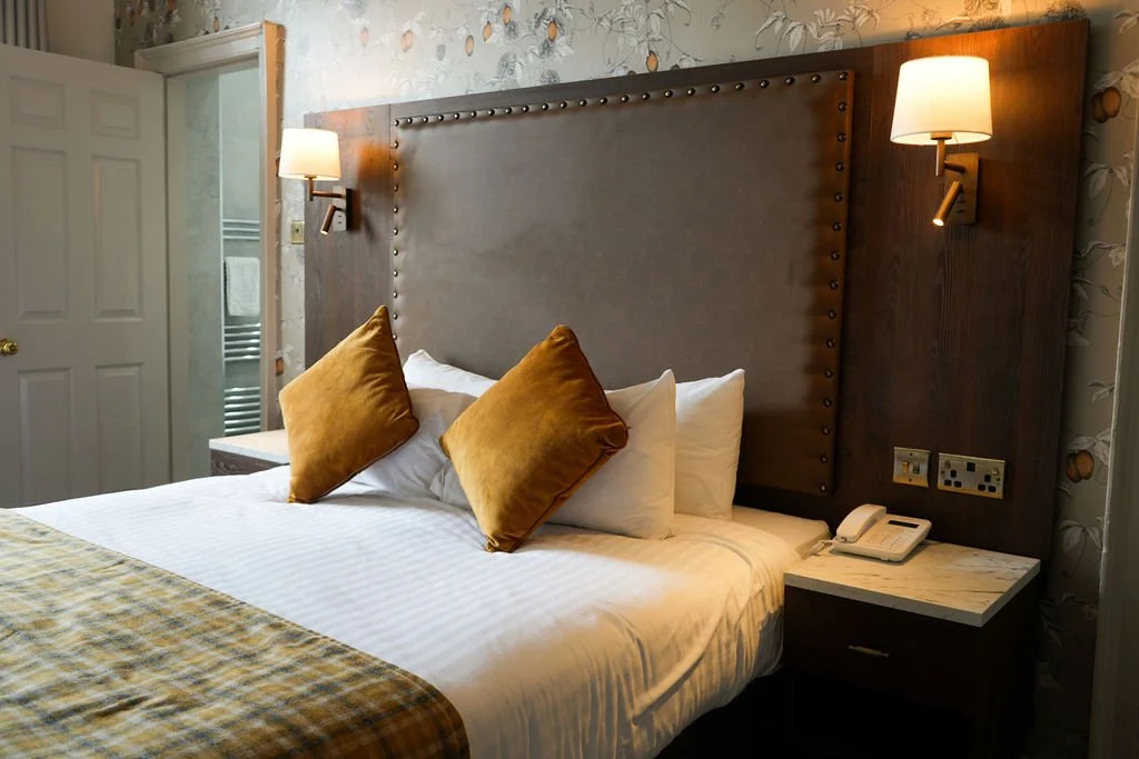 A neatly made bed with white linens, two white pillows, and two gold-colored decorative pillows, in a hotel room with wall-mounted lamps and a marble-topped bedside table with a telephone.