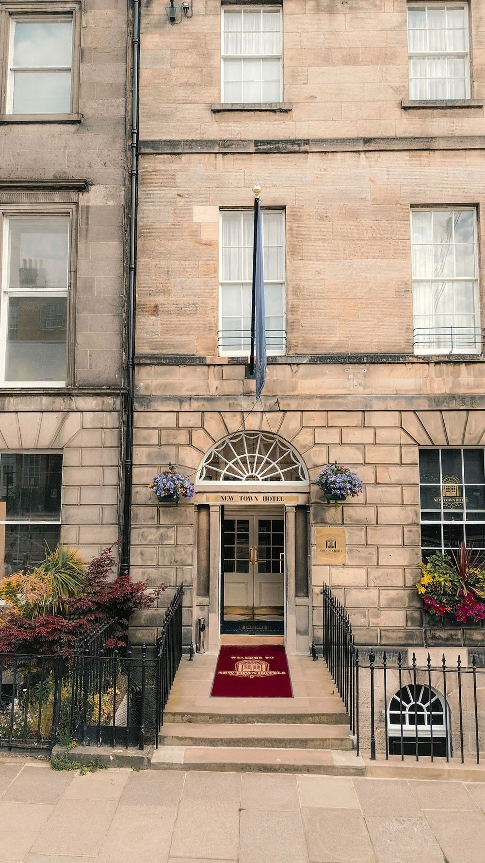 The entrance of the New Town Hotel features steps leading to a glass door, a red carpet with the hotel logo, flower arrangements, and a sign above the door. The building has a stone facade with large windows, and a flag hangs above the entrance.