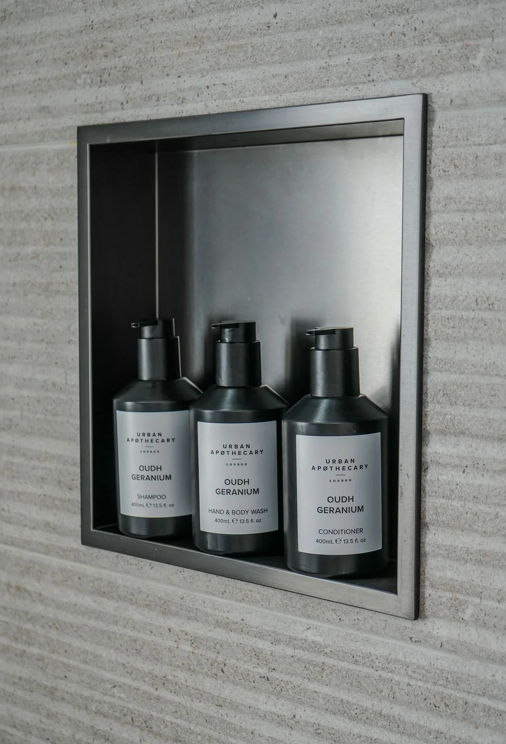 Three black bottles of Urban Apothecary Oudh Geranium toiletries, including shampoo, hand and body wash, and conditioner, displayed in a recessed wall shelf with a metallic border, against a textured wall.
