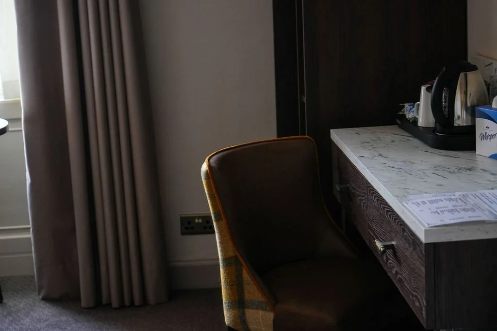 A partially visible hotel room corner with a window curtain, a power outlet, a chair with a plaid fabric cover, a marble countertop, and a tray holding a kettle, cups, and a paper of documents.