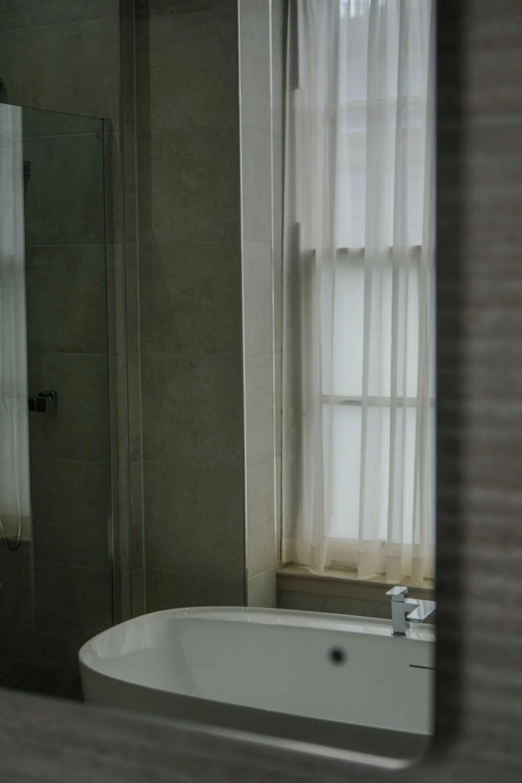 A bathroom with a bathtub and a large window with sheer white curtains, beige tiled walls, and a partial view of a shower area.