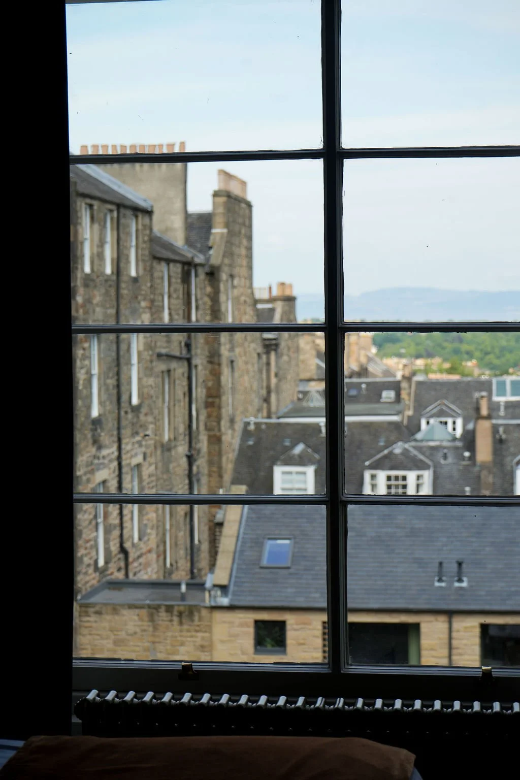 View through a window showing a cityscape with old stone buildings, rooftops, and a distant landscape with hills under a partly cloudy sky.