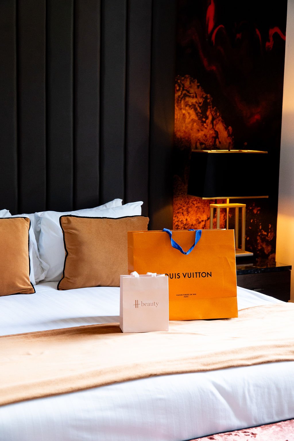 Luxurious hotel bed with beige pillows, two shopping bags from Louis Vuitton and H beauty on the bed, and a black bedside lamp with a gold base, in front of a textured, dark wall.