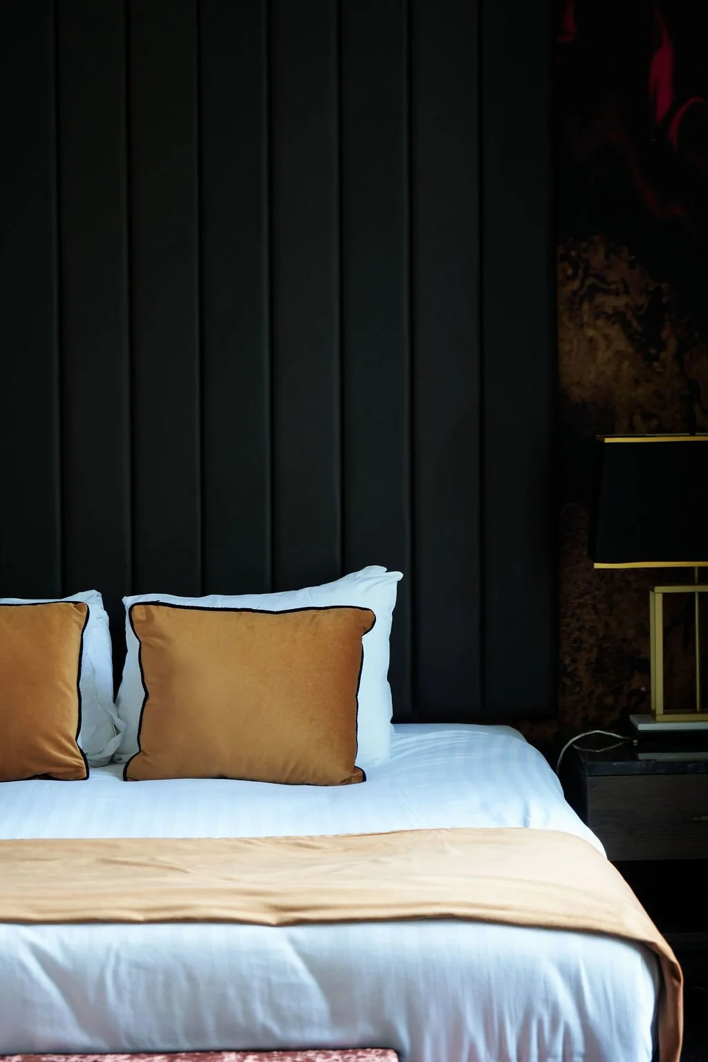 A bed with white sheets, a beige bed runner, and three gold-colored pillows, set against a dark, padded headboard with vertical panels in a dimly lit room. There is a nightstand with a black and gold lamp to the right.
