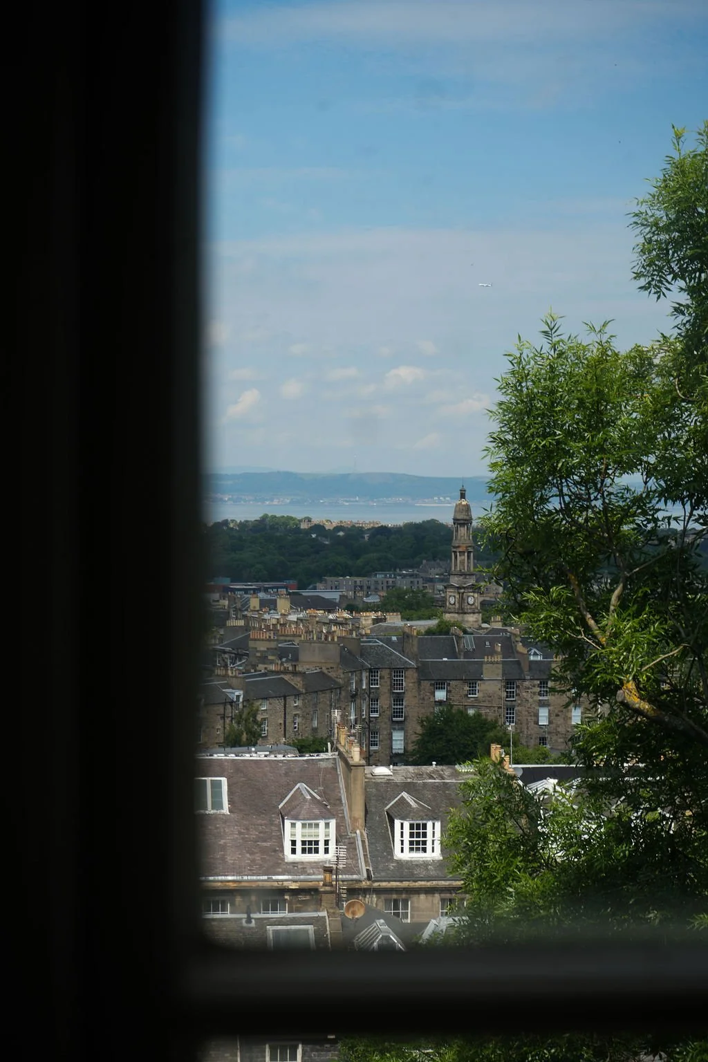 View of a cityscape through a window, showing rooftops, a church steeple, trees, and a distant body of water with hills in the background on a partly cloudy day.