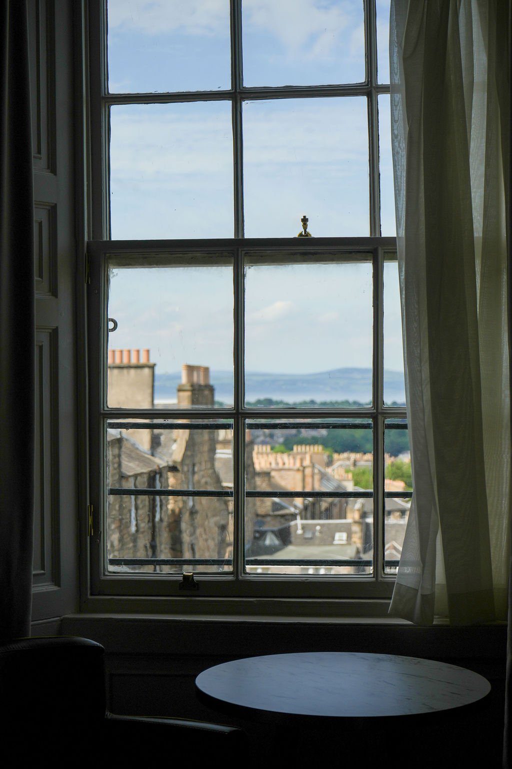 A window with nine panes looking out onto rooftops, chimneys, and a distant body of water with land in the background, partly covered by a curtain.