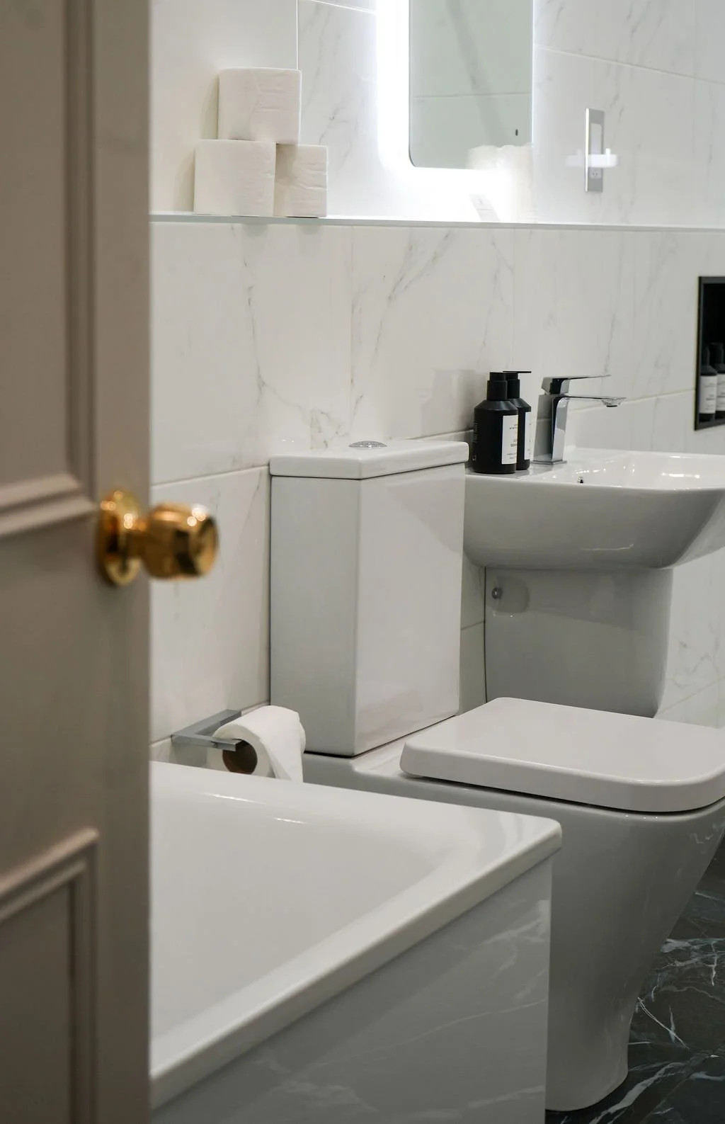 A modern bathroom with marble walls, a toilet, a sink with black soap dispenser, a mirror, and three rolls of toilet paper on shelves.