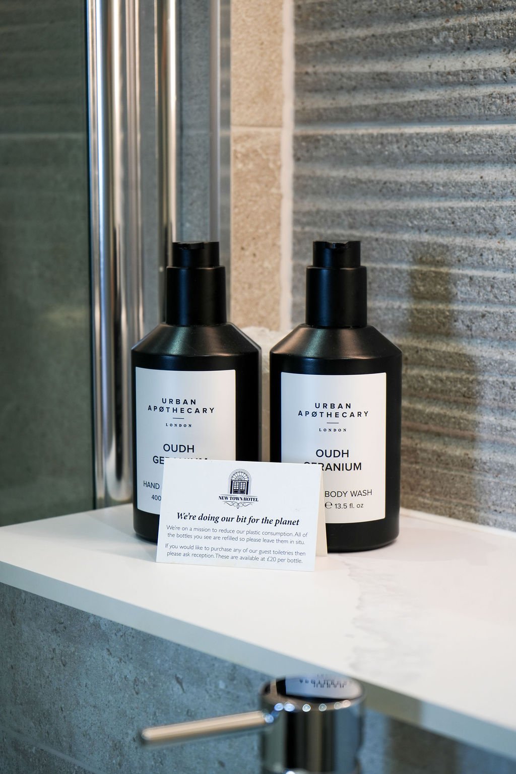 Two black bottles of toiletries, labeled 'Urban Apothecary London' with one labeled 'Oudh Geranium' and the other 'Oudh Titanium', placed on a white shelf with a note in front.