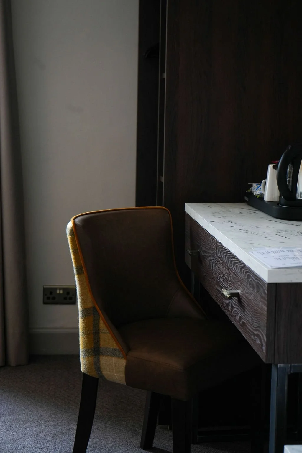 Part of a room with a brown and yellow patterned chair, a white marble-top counter, electrical outlets, and a dark wood cabinet. On the counter are a black kettle, white mugs, and some papers.