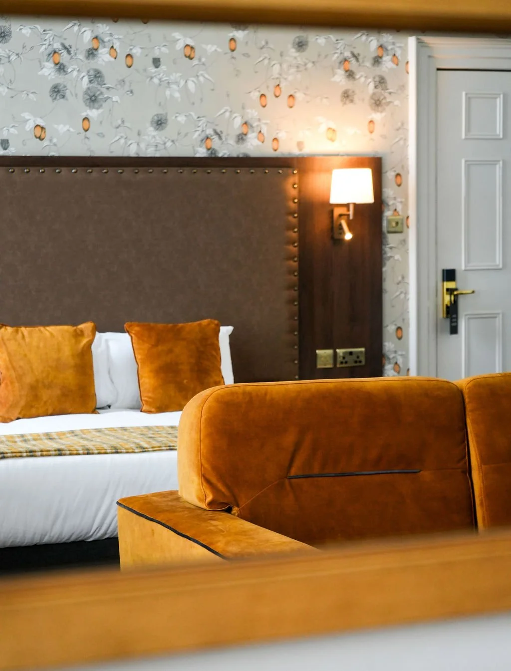 Interior of a hotel room featuring a large upholstered headboard, orange pillows, a bed with a plaid blanket, a wall-mounted lamp, floral wallpaper, and a white door.