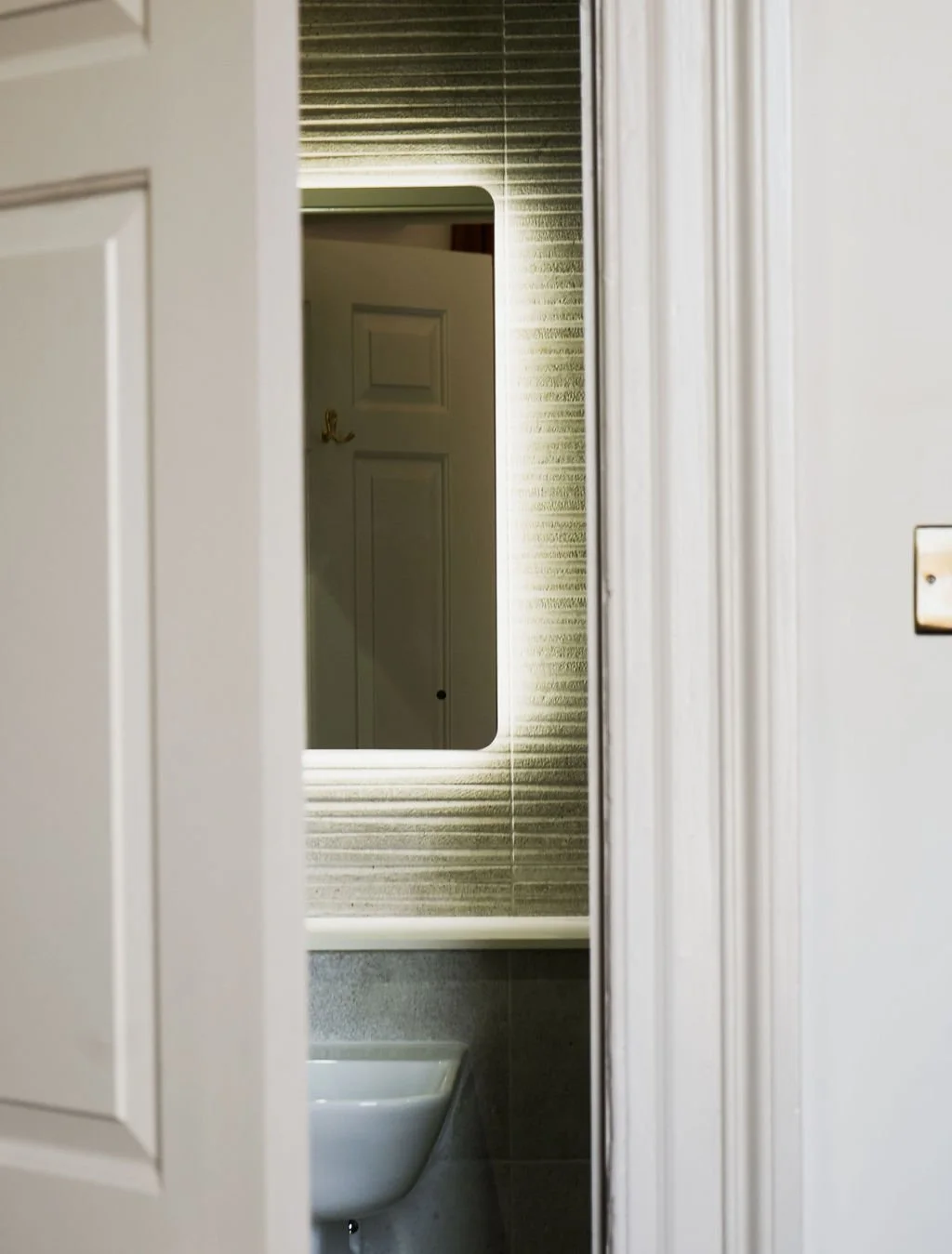 A view through a partially open door reveals a mirror on the wall with a bathroom sink below it. The mirror reflects a closed door in the background.