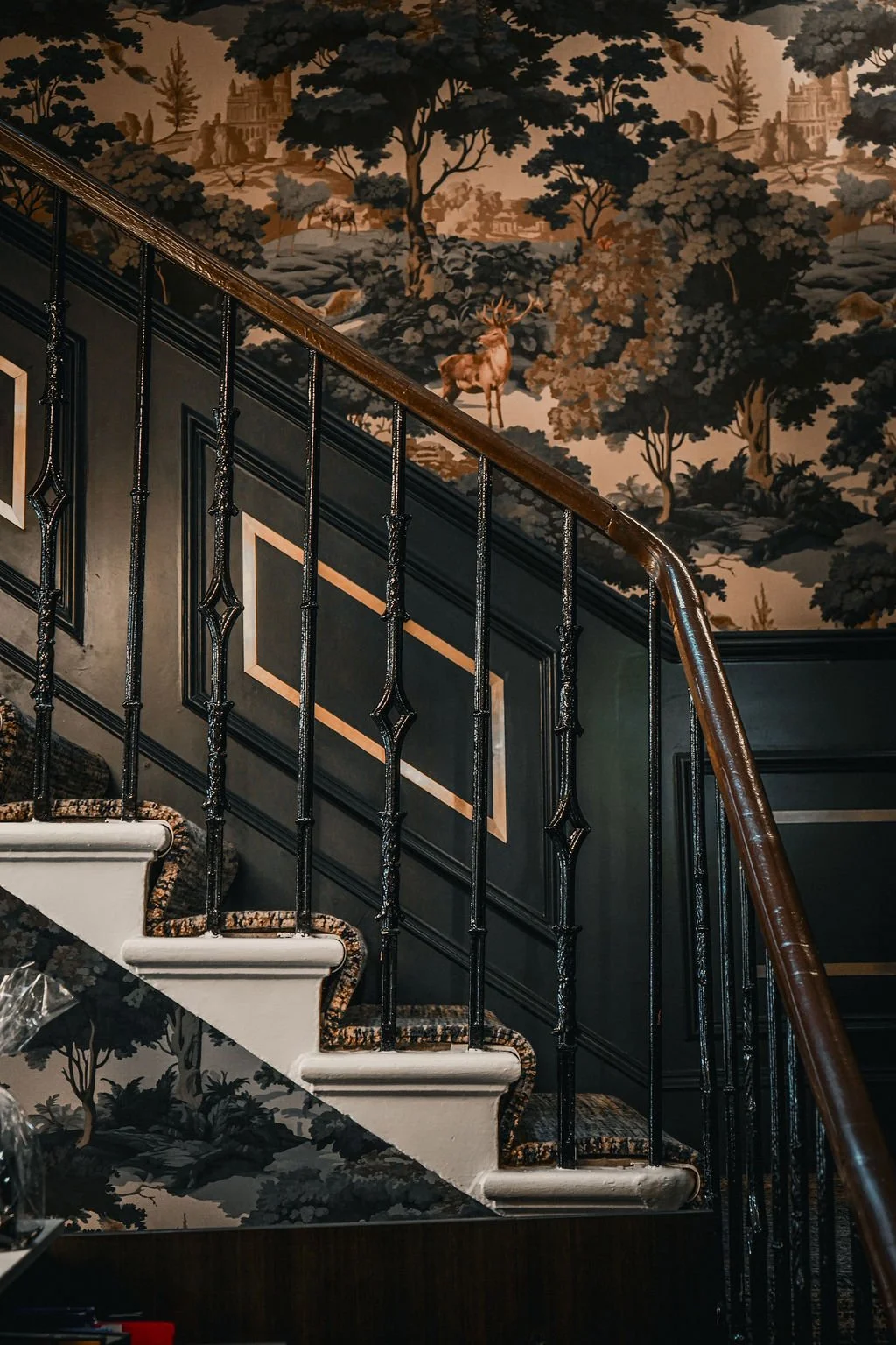 Interior view of a staircase with black ornate iron railings, white steps with a patterned carpet runner, and a wall with gold accents and a scenic wallpaper depicting trees, deer, and distant buildings in a pastoral landscape.