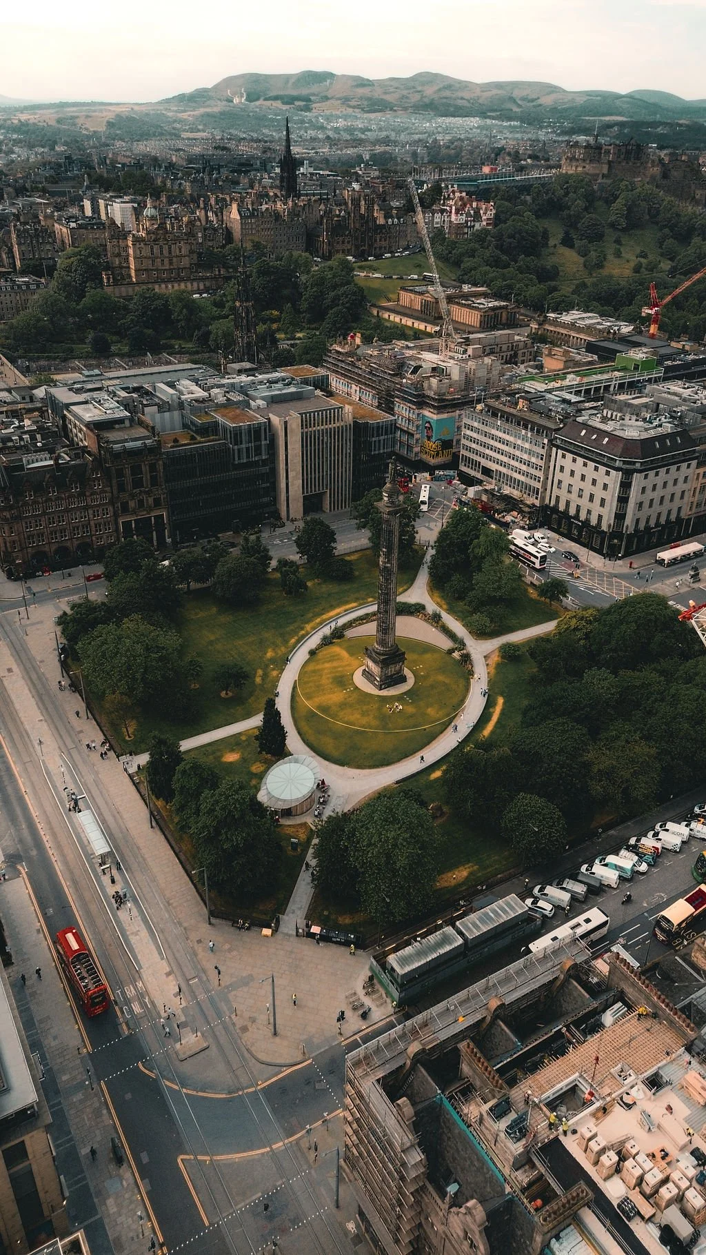 An aerial view of Edinburgh, Scotland, with the Scott Monument prominently in the center, surrounded by trees and nearby buildings, with Edinburgh Castle visible in the distance on a hill.