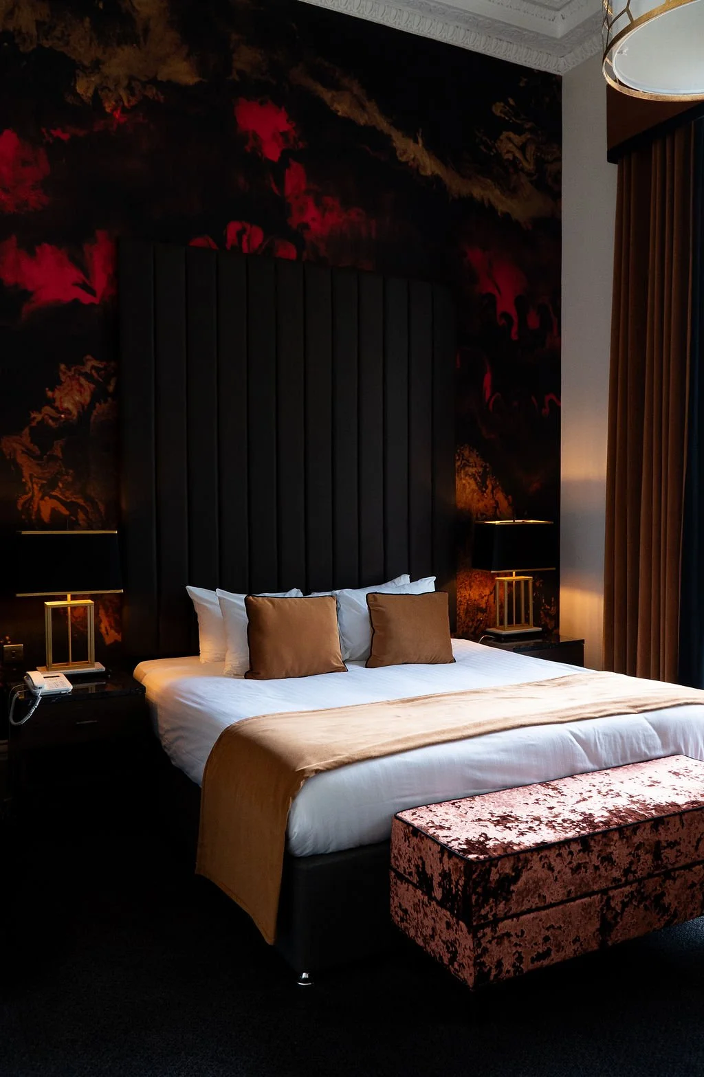 A hotel room with a large bed featuring white sheets, beige pillows, and a beige throw blanket. There is a velvet pink ottoman at the foot of the bed. The wall behind the bed has a dark, textured wallpaper with hints of red and orange. Two black bedside lamps with gold accents are on black nightstands, and there are brown curtains to the right.