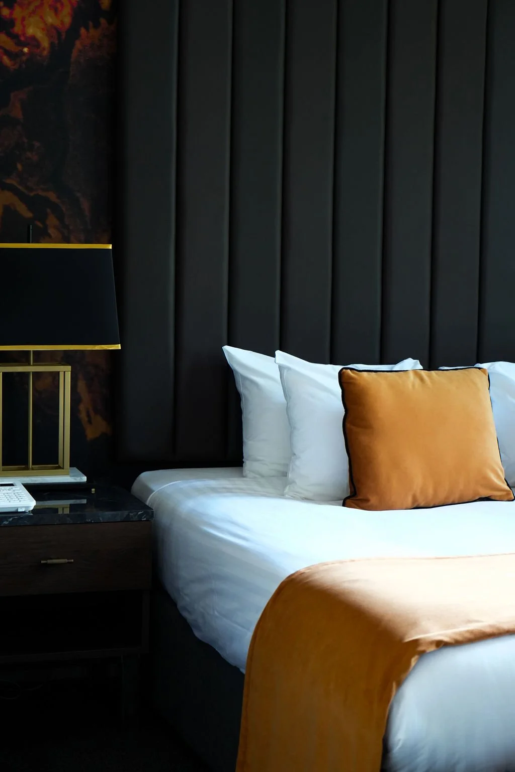 A modern hotel room with a bed that has white sheets, white pillows, and a brown accent pillow. The headboard is tall and upholstered in dark gray fabric. A black and gold table lamp is on a dark wooden nightstand to the left of the bed.
