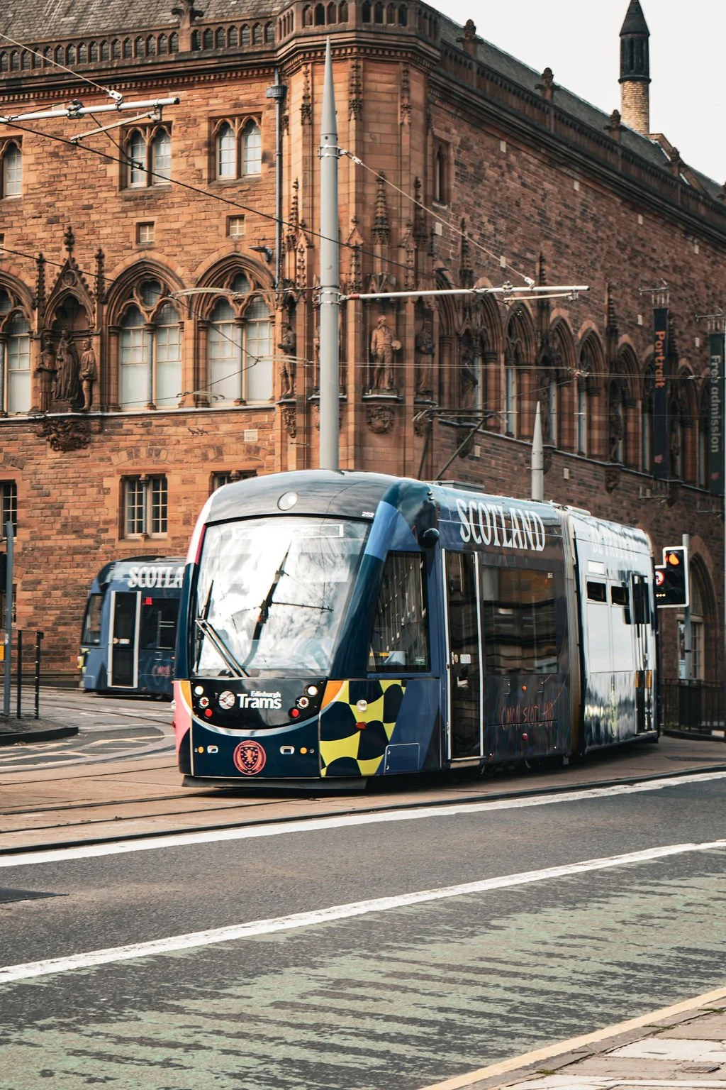 A modern tram with Scotland written on it, traveling past historic red brick buildings with decorative statues and arched windows in an urban setting.
