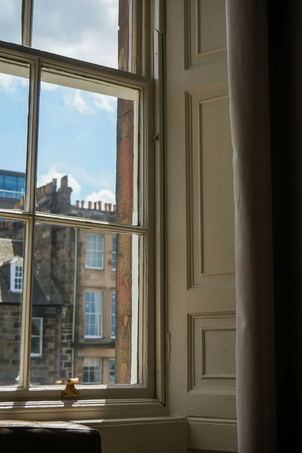 View through a window showing neighboring brick buildings with multiple chimney stacks, blue sky with scattered clouds, and interior part of a window frame with a curtain on the right side.