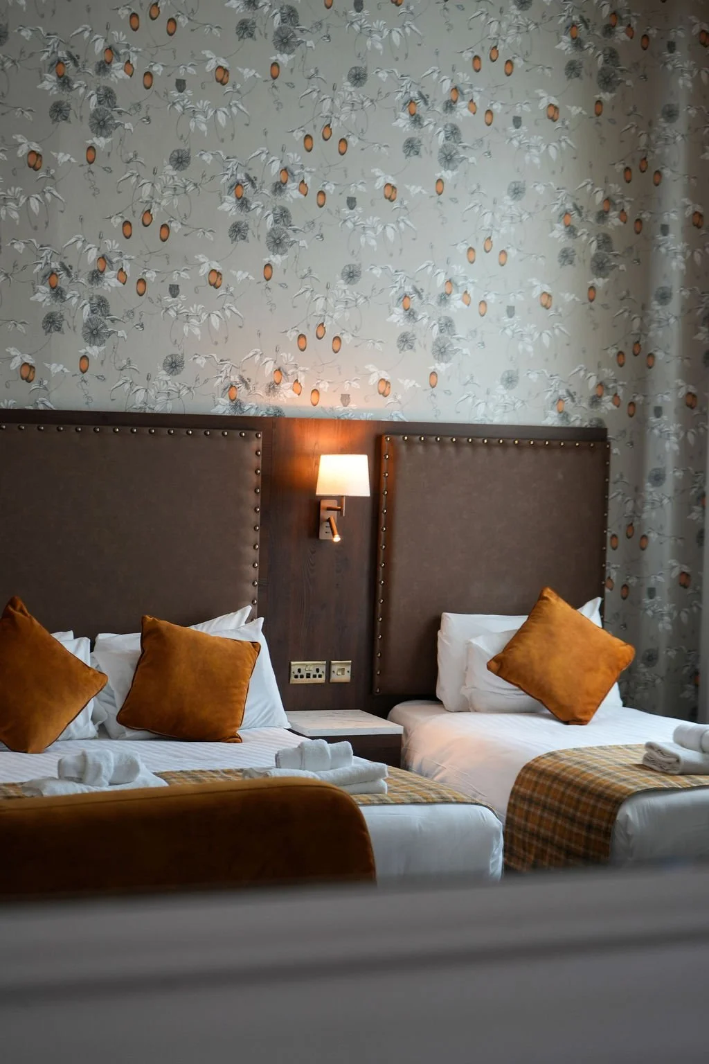 A hotel room with two beds, each with white sheets, brown pillows, and orange plaid bed runners. There is a wall-mounted lamp between the beds and a wall with floral wallpaper featuring fruit patterns.