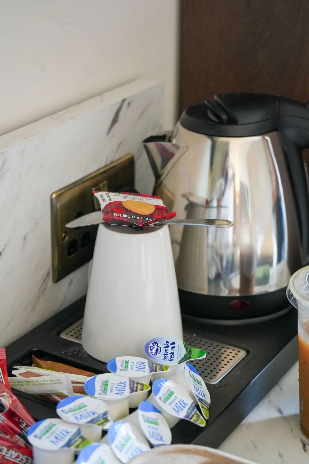 A collection of coffee and creamers, including a small container of vanilla creamer, a small container of milk, and a packet of butter on a tray in front of an electric kettle and a wall socket.