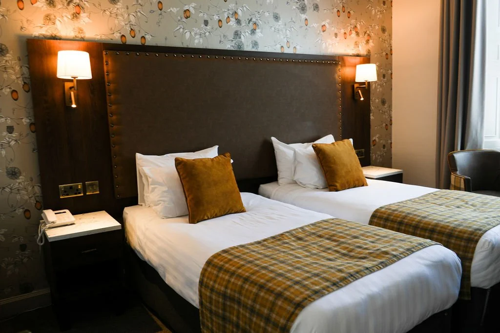 A hotel room with two beds, each with white linens, a honey-colored throw blanket, and a brown pillow. The dark wood headboard spans both beds, with wall-mounted lamps on each side. There are nightstands, one with a telephone, and a chair with a circular back. The wallpaper features a floral pattern with butterflies, and a window with curtains is visible on the right side.