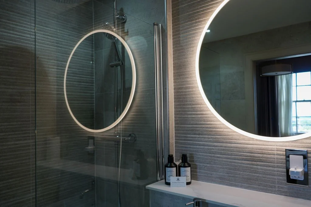 Modern bathroom with a glass shower door, a round illuminated mirror, and toiletries on a shelf.