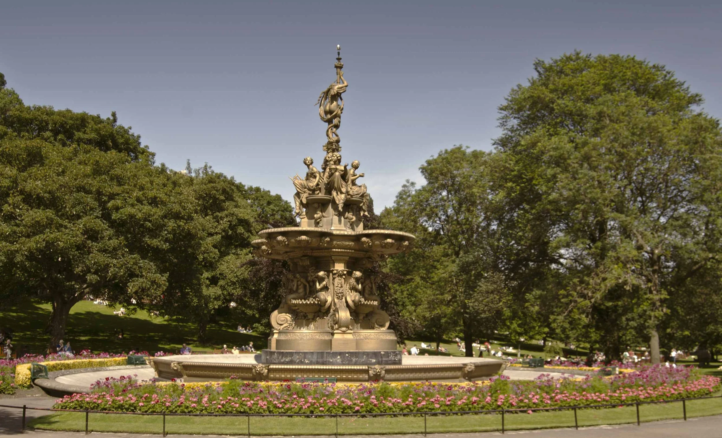 A decorative fountain featuring intricate sculptures of women, with a central column topped by a torch, surrounded by greenery and trees in a park.